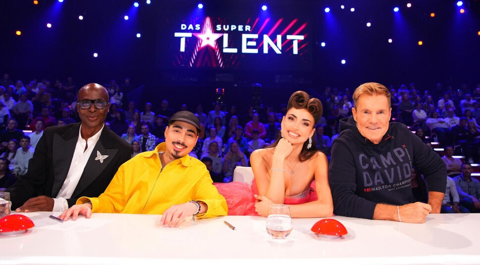 Die Jury von Das Supertalent 2025 sitzt an ihrem Jurypult mit dem Show-Logo und dem Publikum im Hintergrund. Von links nach rechts sind Bruce Darnell in einem eleganten schwarzen Blazer mit Schmetterlingsbrosche, Tony Bauer in einer auffälligen gelben Jacke, Ekaterina Leonova in einem glamourösen pinken Kleid und Dieter Bohlen in einem dunklen Camp David-Hoodie zu sehen. Alle lächeln und wirken gut gelaunt.