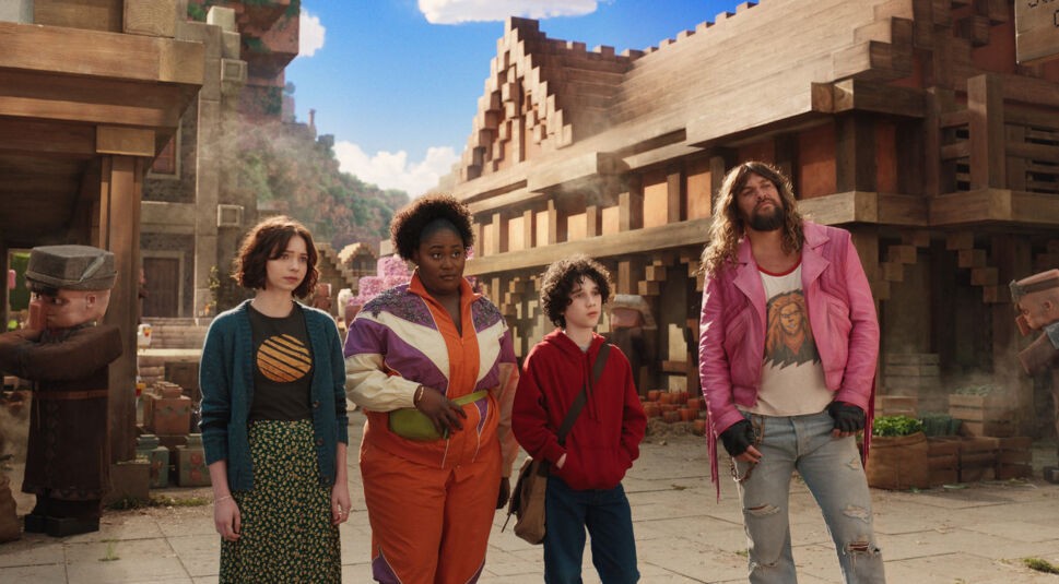 Auf dem Bild aus Ein Minecraft Film stehen Emma Myers als Natalie, Danielle Brooks als Dawn, Sebastian Hansen als Henry und Jason Momoa als Garrett in einem lebendig dargestellten Minecraft-Dorf. Die Umgebung zeigt typische Blockbauten, Dorfbewohner und Marktstände.