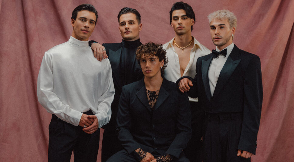 Auf dem Bild zum Elevator Boys Film sind die fünf Mitglieder der Elevator Boys in eleganter Abendgarderobe vor einem rosafarbenen Stoffhintergrund zu sehen. In der Mitte sitzt ein Mitglied in einem schwarzen Anzug mit Spitzen-Details, während die anderen vier in stilvollen Outfits um ihn herumstehen. Ihre Blicke sind ernst und stilbewusst, was eine glamouröse und geheimnisvolle Stimmung vermittelt.