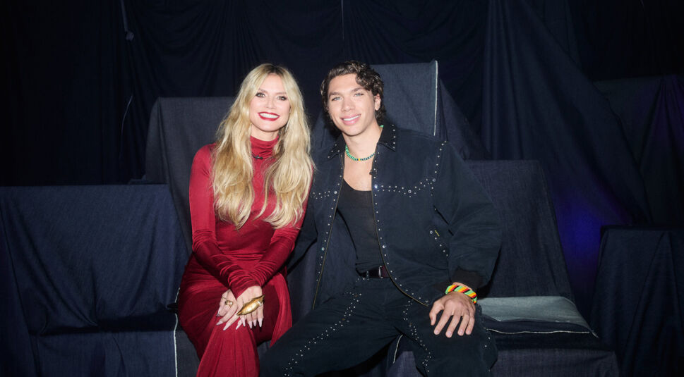 Auf dem Bild zu GNTM 2025 sitzen Heidi Klum und Elias Becker nebeneinander vor dunklem Hintergrund. Heidi trägt ein elegantes, rotes Kleid und lächelt in die Kamera, während Elias in einem schwarzen, mit Nieten besetzten Outfit ebenfalls entspannt lächelt. Die Atmosphäre wirkt freundlich und glamourös – passend zum TV-Format.