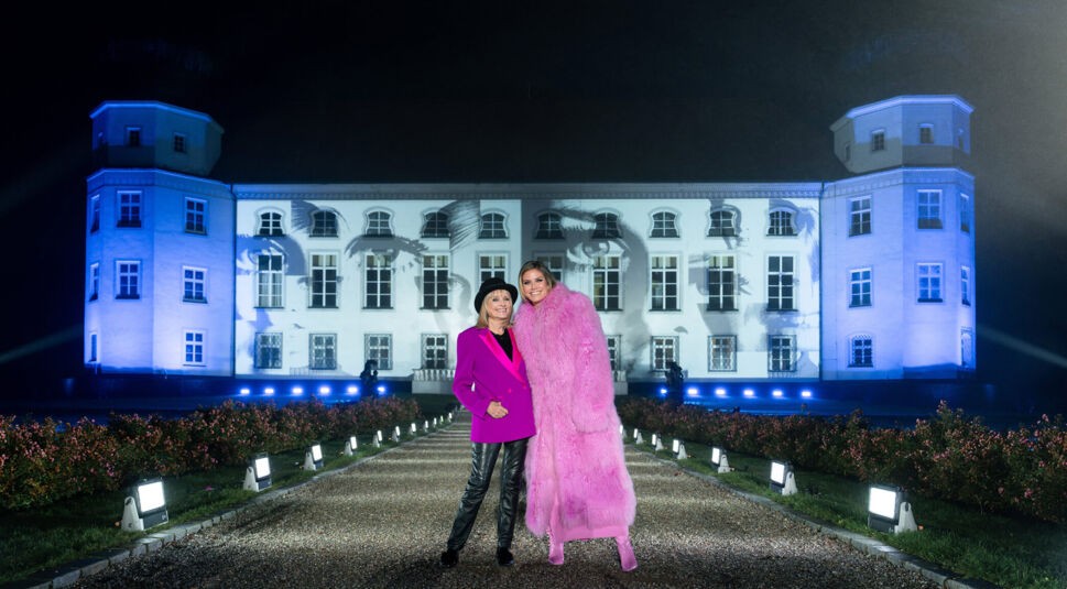 Auf dem Bild zu GNTM 2025 Folge 9 stehen Twiggy und Heidi Klum vor einem eindrucksvoll beleuchteten Schloss. Twiggy trägt einen schwarzen Hut, ein pinkes Sakko und schwarze Lederhosen, während Heidi Klum in einem voluminösen pinken Kunstpelzmantel und passenden Stiefeln strahlt. Im Hintergrund ist eine Projektion mit großen Augen auf die Fassade des Schlosses geworfen, was der Szene eine besondere Atmosphäre verleiht.