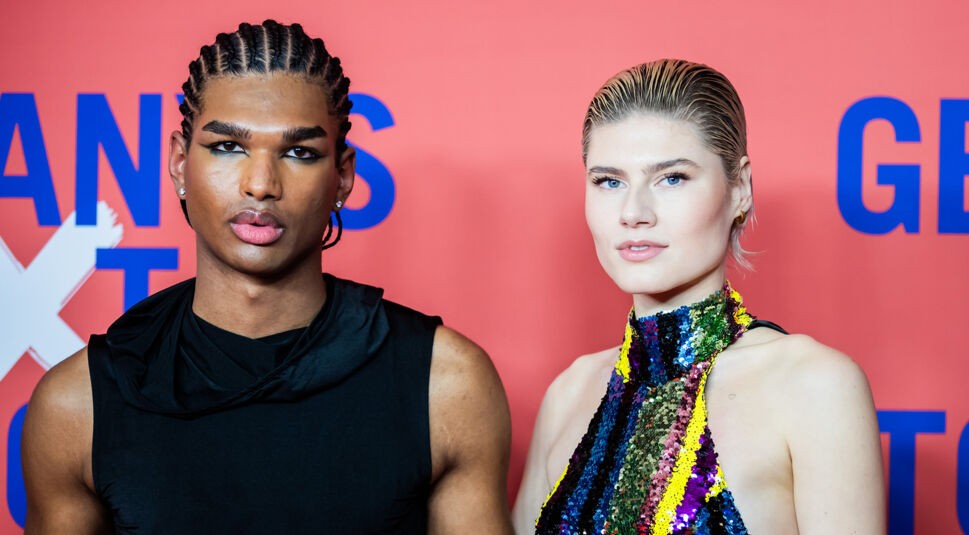 Auf dem Bild in dem Artikel zu den GNTM-Gewinner:innen sind Jermaine und Lea vor einem roten Hintergrund mit GNTM-Schriftzug zu sehen. Jermaine trägt ein schwarzes ärmelloses Oberteil und auffälliges Augen-Make-up. Lea trägt ein bunt glitzerndes Neckholder-Kleid mit zurückgegeltem Haar. Beide blicken selbstbewusst in die Kamera.