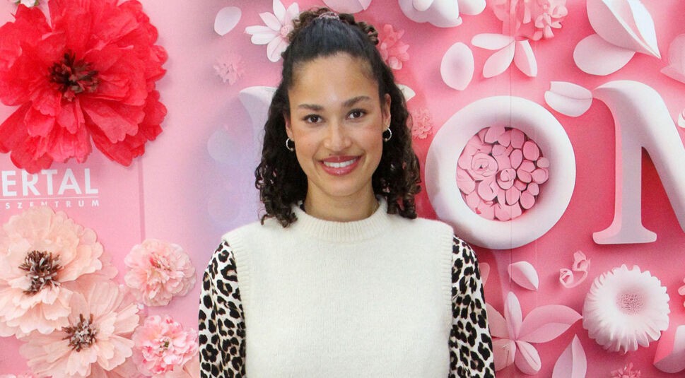 Auf dem Bild in dem Artikel zu den GNTM-Gewinner:innen ist Lovelyn Enebechi vor einem rosafarbenen Hintergrund mit großen Papierblumen und Dekoelementen zu sehen. Sie trägt einen weißen Pullunder über einem langärmligen Oberteil mit Leopardenmuster. Ihr lockiges Haar ist halb hochgesteckt, und sie lächelt freundlich in die Kamera.