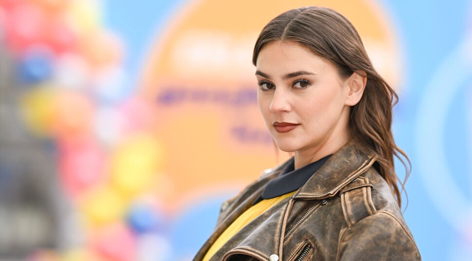 Auf dem Bild in dem Artikel zu den GNTM-Gewinner:innen ist Stefanie Giesinger vor einem bunten, unscharfen Hintergrund mit Orange-, Blau- und Gelbtönen zu sehen. Sie trägt eine abgewetzte Lederjacke über einem gelben Oberteil mit schwarzem Kragen. Ihr braunes Haar fällt offen über die Schultern, und sie blickt ernst und selbstsicher in die Kamera.