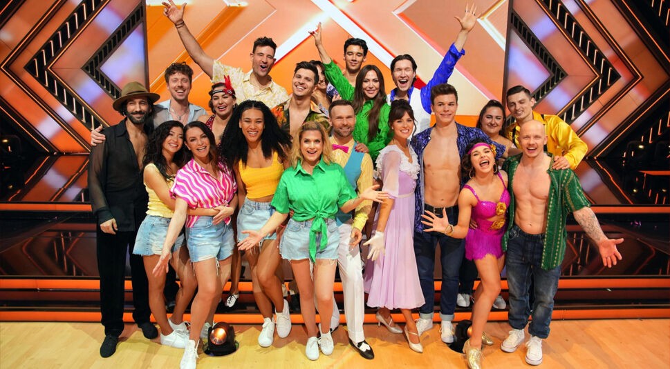 Auf dem Gruppenbild zur Let's Dance 2025 Show 5 stehen mehrere strahlende Tanzpaare in bunten Outfits auf der Studiobühne. Die Stimmung ist ausgelassen und fröhlich, alle lachen in die Kamera. Die Bühne im Hintergrund leuchtet in Orange- und Goldtönen. Die Tanzpaare posieren eng nebeneinander und zeigen sich bereit für ihren Auftritt bei Let's Dance 2025 Show 5.