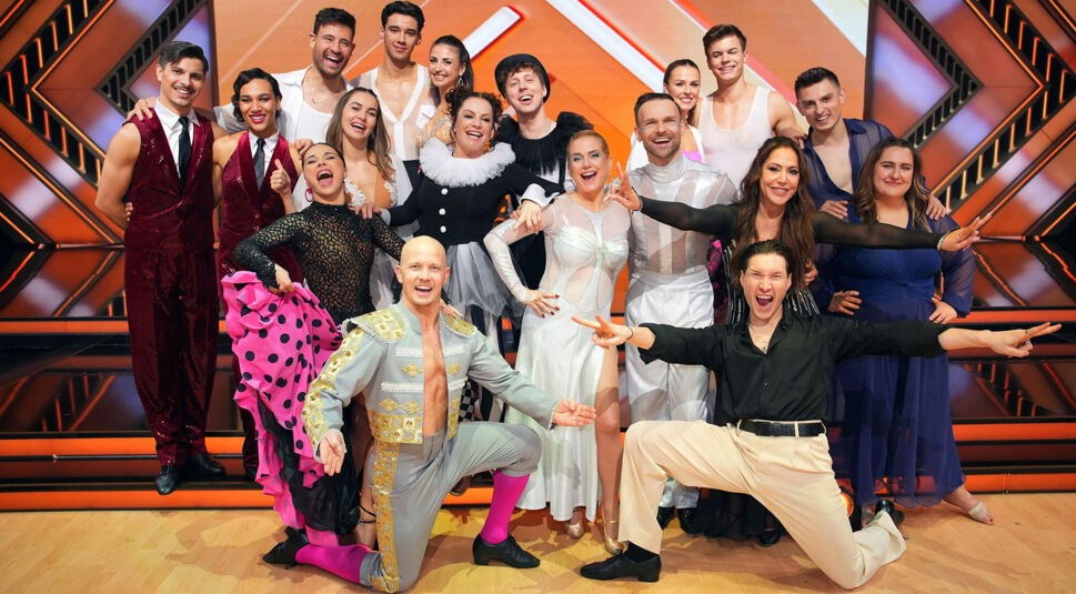 Auf dem Bild zur Let's Dance 2025 Show 6 ist die gesamte Tanz-Crew in bunten, auffälligen Kostümen vor einem leuchtenden Show-Hintergrund zu sehen. Die Gruppe strahlt vor Freude, viele lachen und posieren ausgelassen. In der ersten Reihe knien zwei Tänzer:innen mit ausdrucksstarker Körpersprache, während die restlichen Teilnehmenden eng zusammenstehen. Die Stimmung ist energiegeladen und feierlich.
