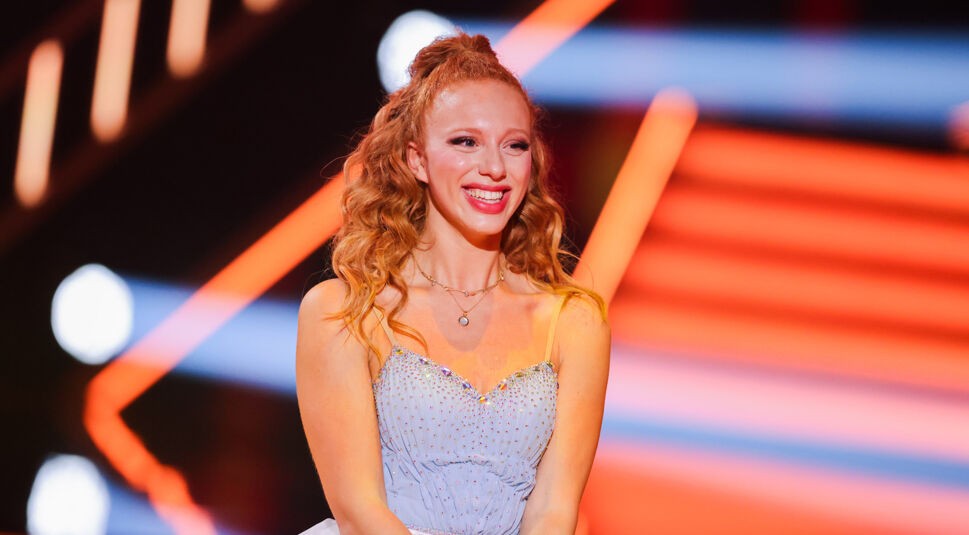 Auf dem Bild in dem Artikel zu den Let's Dance Gewinner:innen ist Anna Ermakova zu sehen. Sie trägt ein elegantes, hellblaues Kleid mit funkelnden Verzierungen und hat ihre lockigen, rötlichen Haare halboffen gestylt. Mit einem strahlenden Lächeln steht sie auf der Bühne, umgeben von leuchtenden, orangefarbenen Lichteffekten, die eine festliche und dynamische Atmosphäre schaffen.