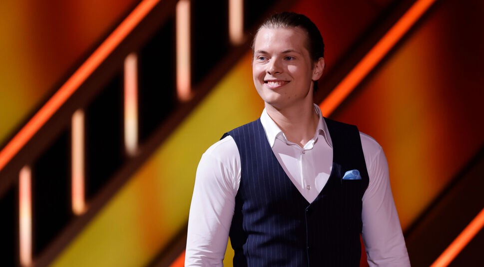 Auf dem Bild in dem Artikel zu den Let's Dance Gewinner:innen ist Gabriel Kelly zu sehen. Er trägt ein elegantes Outfit mit einem dunkelblauen Westenanzug, einem weißen Hemd und einem hellblauen Einstecktuch. Mit einem zufriedenen Lächeln blickt er zur Seite, während der Hintergrund mit warmen orange-gelben Lichteffekten eine dynamische Bühnenszene schafft.