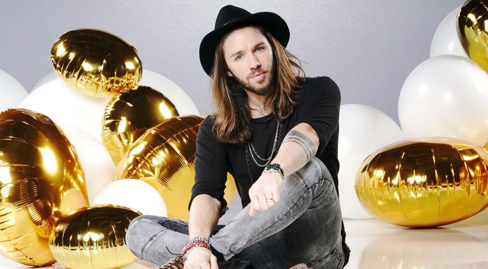 Auf dem Bild in dem Artikel zu den Let's Dance Gewinner:innen ist Gil Ofarim zu sehen. Er sitzt entspannt auf dem Boden, umgeben von goldenen und weißen Luftballons. Er trägt einen schwarzen Hut, ein schwarzes Shirt und graue Jeans. Seine langen Haare und Accessoires wie Armbänder und Ketten unterstreichen seinen lässigen Look. Sein Blick ist direkt und ausdrucksstark in die Kamera gerichtet.