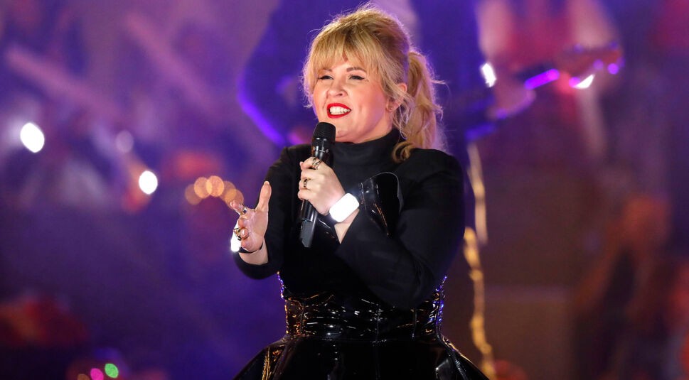 Auf dem Bild in dem Artikel zu den Let's Dance Gewinner:innen ist Maite Kelly zu sehen. Sie steht auf der Bühne und hält ein Mikrofon in der Hand, während sie mit ausdrucksstarker Gestik performt. Sie trägt ein elegantes schwarzes Outfit mit glänzenden Details und ihre blonde Frisur ist in einem lockeren Zopf gestylt. Der Hintergrund ist in dunklen, stimmungsvollen Farben gehalten, mit Lichteffekten und verschwommenen Silhouetten des Publikums.