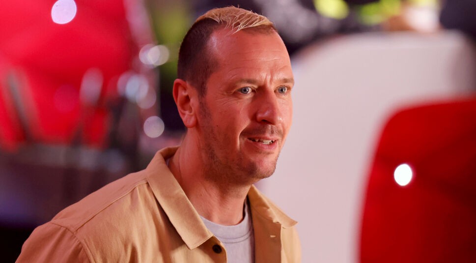 Auf dem Bild in dem Artikel zu den Let's Dance Gewinner:innen ist Pascal Hens zu sehen. Er trägt eine beige Jacke über einem weißen Shirt und hat eine Kurzhaarfrisur mit blond gefärbter Spitze. Sein Gesichtsausdruck wirkt freundlich und aufgeschlossen, während er leicht zur Seite blickt. Der Hintergrund ist unscharf und enthält rote und helle Farbakzente, die eine lebendige Atmosphäre schaffen.