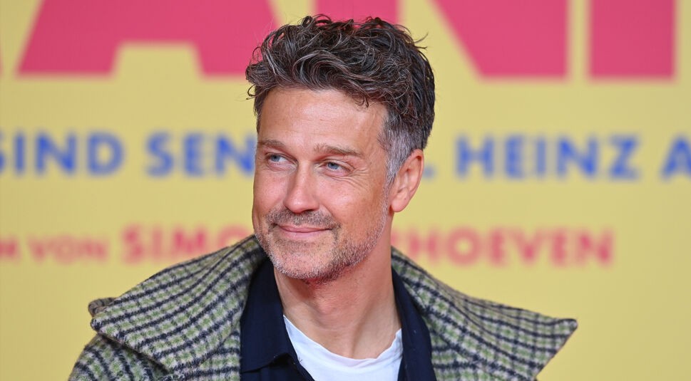 Auf dem Bild in dem Artikel zu den Let's Dance Gewinner:innen ist Wayne Carpendale zu sehen. Er trägt ein lässiges Outfit mit einem weißen Shirt, einem dunklen Hemd und einer karierten Jacke. Sein Blick ist leicht zur Seite gerichtet, begleitet von einem dezenten Lächeln. Der Hintergrund zeigt eine bunte Schrift auf gelbem Untergrund, die auf ein Event oder eine Premiere hinweist.