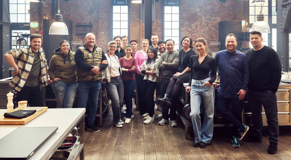 Auf dem Bild zur Sendung Mälzers Meisterklasse VOX steht eine Gruppe von Teilnehmer:innen gemeinsam mit Tim Mälzer in einer modernen Studioküche. 