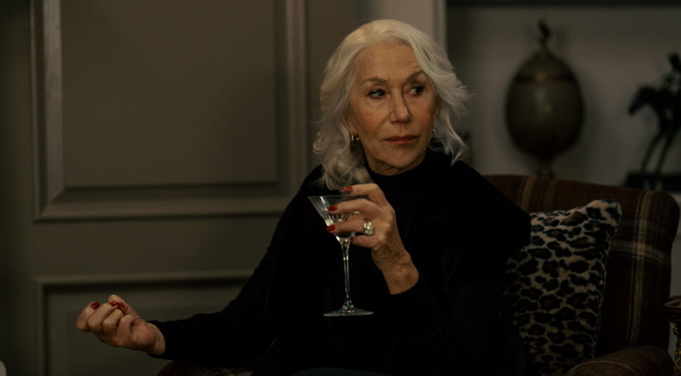 Auf dem Bild zur Mobland Serie ist Helen Mirren als Maeve Harrigan zu sehen. Sie sitzt in einem eleganten, gedämpft beleuchteten Raum auf einem Sessel mit einem gemusterten Kissen. Gekleidet in Schwarz, hält sie ein Martini-Glas in der Hand und trägt roten Nagellack. Ihr silbernes Haar fällt in sanften Wellen, während sie mit einem nachdenklichen Ausdruck zur Seite blickt.