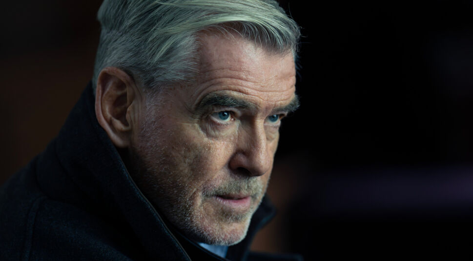 Auf dem Bild zur Mobland Serie ist Pierce Brosnan als Conrad Harrigan zu sehen. Er trägt einen dunklen Mantel mit hochgeschlagenem Kragen und hat einen intensiven, durchdringenden Blick. Sein silbergraues Haar ist zurückgekämmt, und sein Gesicht zeigt einen leichten Bartschatten. Die düstere Beleuchtung und der verschwommene Hintergrund unterstreichen die ernste, geheimnisvolle Atmosphäre der Szene.