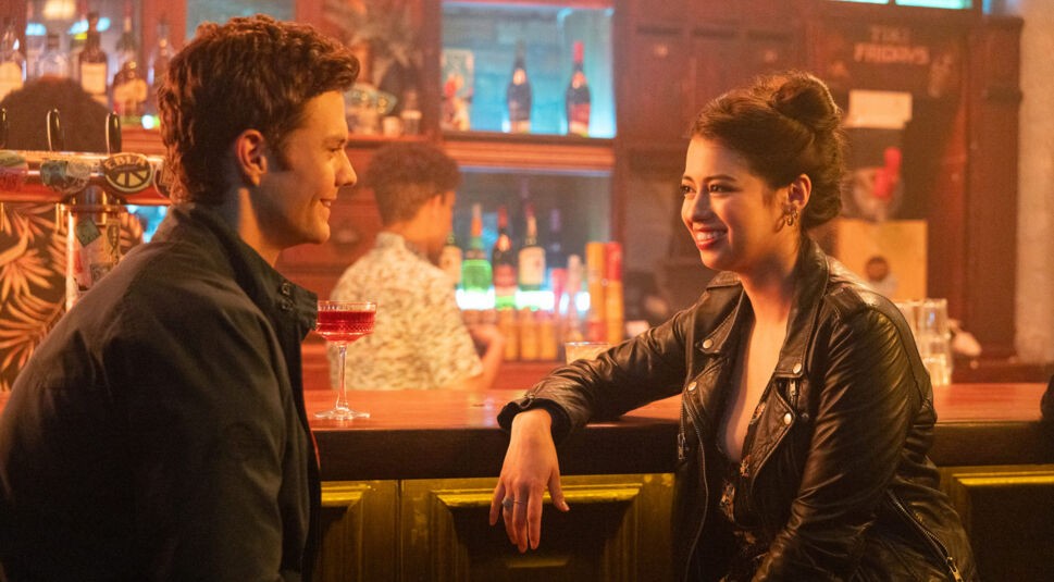 Auf dem Bild aus Mr. No Pain Kritik sind Jack Quaid als Nate und Amber Midthunder als Sherry in einer Bar zu sehen. Die beiden sitzen sich lächelnd gegenüber, während ein roter Cocktail auf dem Tresen steht. Sherry trägt eine schwarze Lederjacke und lehnt entspannt an der Theke, während Nate in dunkler Kleidung interessiert zuhört. Die warme Beleuchtung und die lebendige Atmosphäre deuten auf eine lockere, charmante Unterhaltung zwischen den Charakteren hin