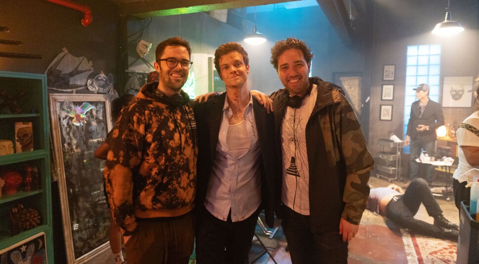 Auf dem Bild aus der Mr. No Pain Kritik sind Regisseur Robert Olsen, Hauptdarsteller Jack Quaid als Nate und Regisseur Dan Berk am Filmset zu sehen. Jack Quaid trägt ein blutverschmiertes Hemd und ein Unterhemd, während er entspannt zwischen den beiden lachenden Regisseuren steht. Die Szenerie im Hintergrund wirkt chaotisch, mit einem bewusstlosen Mann auf dem Boden und einer düsteren, neonbeleuchteten Atmosphäre, die die Actionfilm-Stimmung unterstreicht.