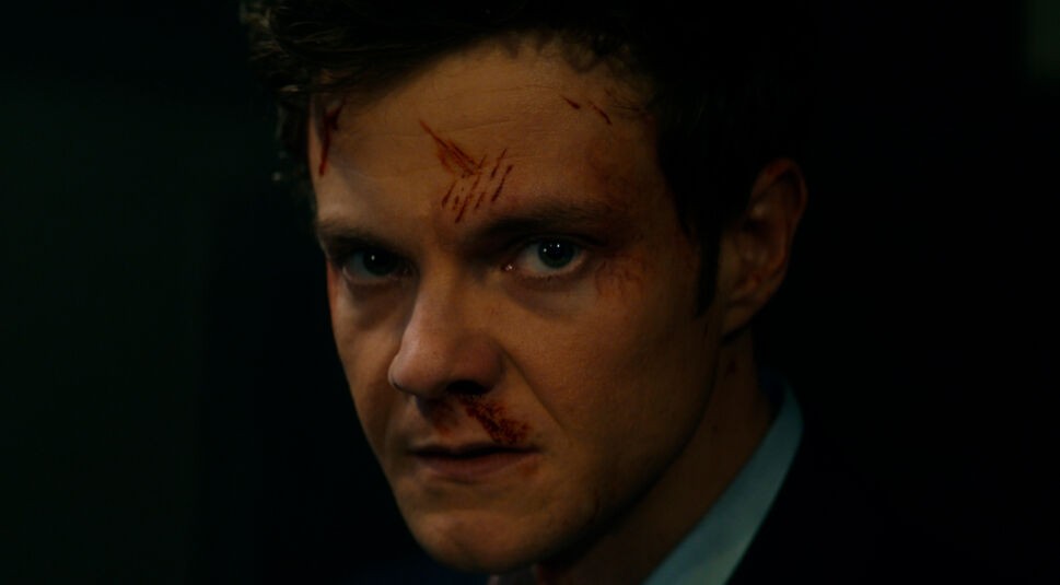 Auf dem Bild aus Mr. No Pain Kritik ist Jack Quaid als Nathan Cain in einer intensiven Nahaufnahme zu sehen. Sein Gesicht ist mit frischen Kratzern und Blutspuren gezeichnet, während sein entschlossener, fast bedrohlicher Blick direkt in die Kamera gerichtet ist. Die düstere Beleuchtung und der starke Schattenwurf verstärken die angespannte und dramatische Atmosphäre der Szene.