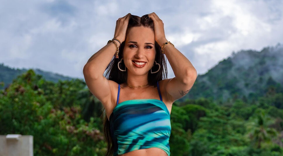 Auf dem Bild zu Princess Charming 2025 ist nessiontour zu sehen, wie sie strahlend in die Kamera lächelt. Sie trägt ein blau-grünes Top mit Spaghettiträgern und kombiniert es mit goldenen Accessoires, darunter herzförmige Ohrringe und Armbänder. Mit beiden Händen greift sie in ihr dunkles Haar. Der Hintergrund zeigt eine tropische Landschaft mit grünen Bergen und bewölktem Himmel, was eine entspannte und abenteuerliche Atmosphäre vermittelt.