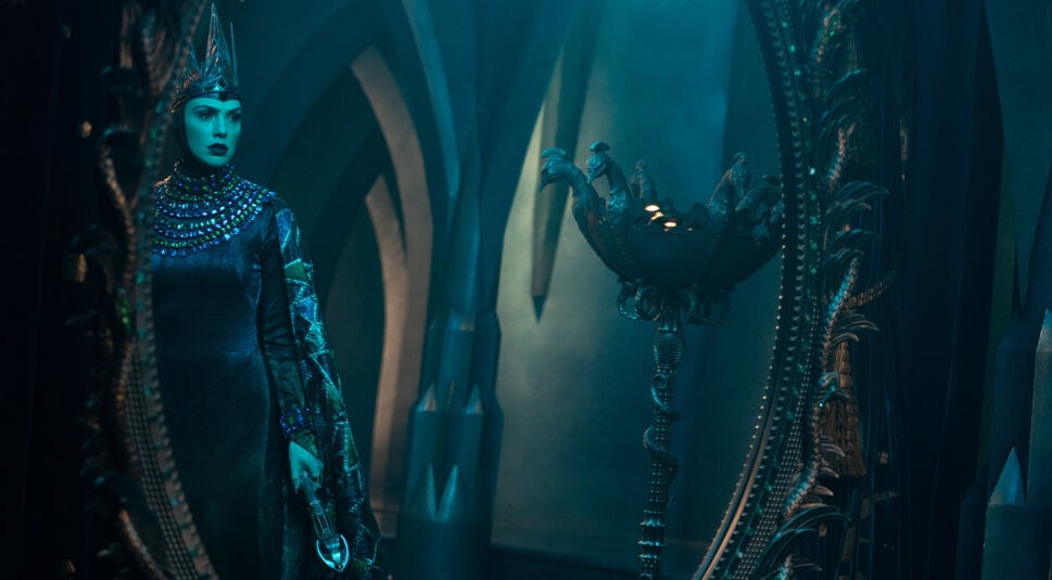 Auf dem Bild aus Schneewittchen (2025) ist Gal Gadot als böse Königin in einem düsteren, imposanten Thronsaal zu sehen. Sie trägt eine schwarze, glänzende Robe mit aufwendigem Edelsteinbesatz und eine spitze Krone. Ihr ausdrucksstarker Blick spiegelt Macht und Bedrohlichkeit wider. Die Szene ist in kühlem, blaugrünem Licht gehalten und hebt sich stilistisch deutlich vom klassischen Animationsstil von Schneewittchen und die sieben Zwerge (1937) ab, was den visuellen Kontrast zwischen beiden Versionen verdeutlicht.