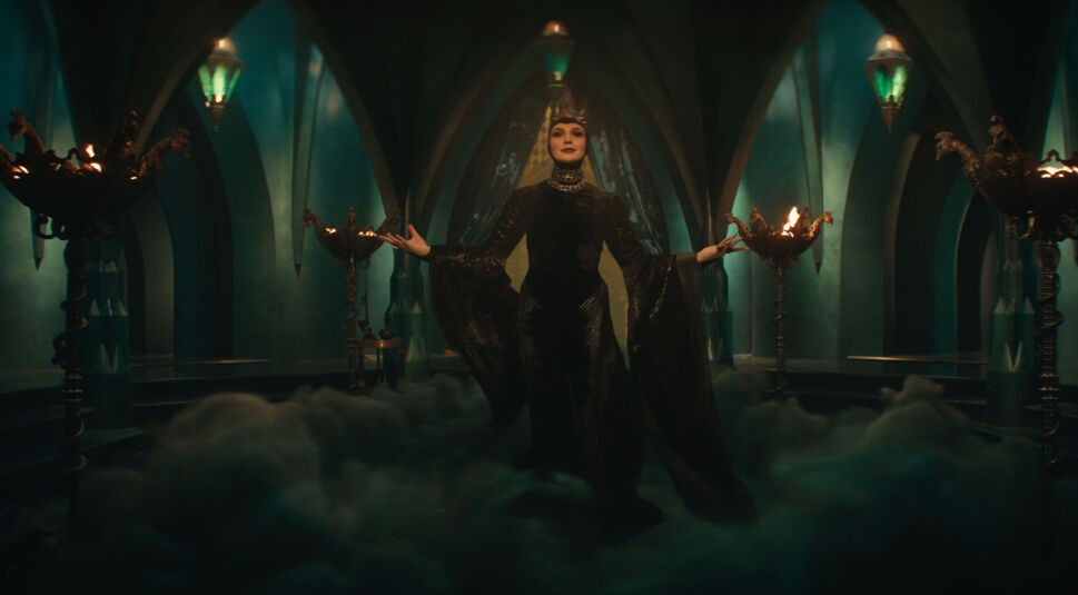 Auf dem Bild in dem Artikel zu der Schneewittchen-Filmkritik ist die böse Königin, gespielt von Gal Gadot, in einer imposanten Pose zu sehen. Sie trägt ein elegantes, schwarzes Gewand mit weit ausladenden Ärmeln und eine verzierte Krone. Ihre Arme sind ausgebreitet, während sie in einem düsteren, grünlich beleuchteten Raum mit Bögen und Feuerschalen steht. Mystischer Nebel am Boden verstärkt die bedrohliche und magische Atmosphäre der Szene.