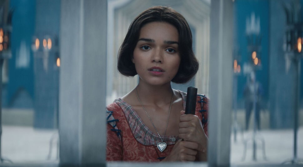 Auf dem Bild in dem Artikel zu der Schneewittchen-Filmkritik ist Schneewittchen, gespielt von Rachel Zegler, in einer ernsten Szene zu sehen. Sie trägt ein schlichteres Kleid in warmen Tönen und hält einen Besenstiel fest umklammert. Ihr Blick wirkt nachdenklich und leicht besorgt, während sie durch eine vergitterte Öffnung schaut. Der Hintergrund zeigt eine elegante, blau gehaltene Schlosskulisse mit Kerzenleuchtern, was die märchenhafte Atmosphäre unterstreicht.