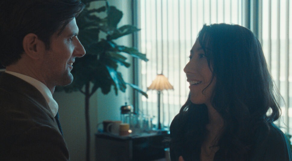 Auf dem Bild aus Severance Staffel 2 sind Adam Scott als Mark Scout und Dichen Lachman als Gemma bzw. Casey in einem warm beleuchteten Büro zu sehen. Die beiden stehen sich nahe und lächeln einander an, was auf eine emotionale oder bedeutungsvolle Begegnung hindeutet. Im Hintergrund sind eine Lampe, Pflanzen und eine Kaffeestation zu erkennen, die eine gemütliche Atmosphäre schaffen. Severance Staffel 2 Ende erklärt – diese Szene könnte auf eine entscheidende Enthüllung rund um Marks Vergangenheit und Gemmas wahre Identität hinweisen.