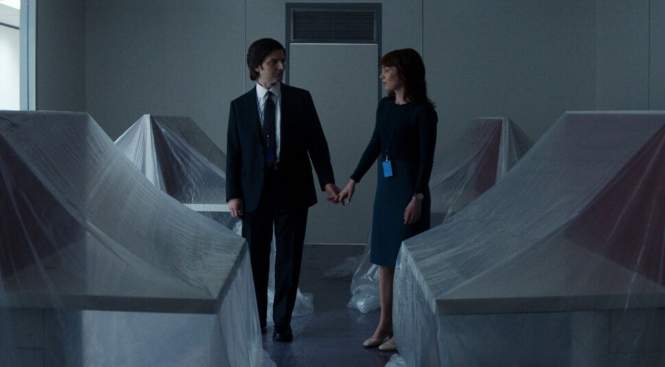 Auf dem Bild aus Severance Staffel 2 sind Adam Scott als Mark Scout und Britt Lower als Helly Riggs in einem düsteren, verlassen wirkenden Raum zu sehen. Die beiden stehen zwischen mit Plastikfolie abgedeckten Schreibtischen und halten sich an den Händen, während sie sich ernst und nachdenklich ansehen. Die sterile, bedrückende Atmosphäre deutet auf eine bedeutende Enthüllung oder einen Wendepunkt in der Handlung hin. Severance Staffel 2 Ende erklärt – diese Szene könnte ein Schlüsselmoment für Marks und Hellys Beziehung innerhalb von Lumon Industries sein.