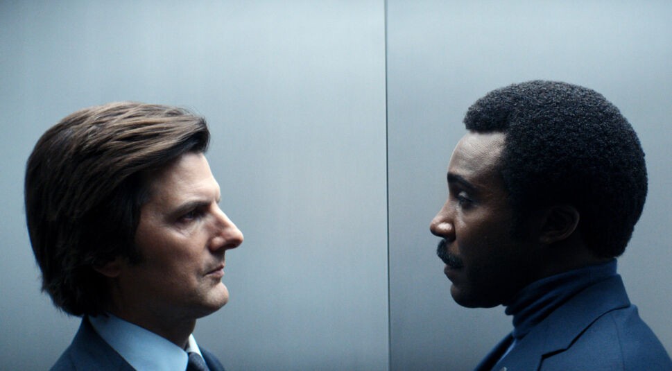 Auf dem Bild aus Severance Staffel 2 stehen sich Adam Scott als Mark Scout und Tramell Tillman als Seth Milchick in einem engen, kühlen Raum gegenüber, vermutlich einem Aufzug. Beide blicken sich mit ernsten, angespannten Gesichtsausdrücken direkt in die Augen. Mark trägt einen dunklen Anzug mit Hemd, während Milchick in einem blauen Rollkragenpullover und Blazer zu sehen ist. Severance Staffel 2 Ende erklärt – diese Szene deutet auf eine bedeutende Konfrontation zwischen den beiden Charakteren hin.