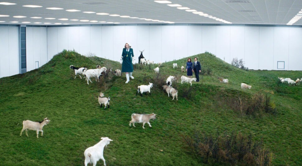 Auf dem Bild aus Severance Staffel 2 sind Gwendoline Christie, Adam Scott als Mark Scout und Britt Lower als Helly Riggs in einer surrealen Szene zu sehen. In einem großen, künstlich beleuchteten Raum erstreckt sich eine grüne Hügellandschaft, auf der zahlreiche Ziegen umherlaufen. Gwendoline Christie trägt einen langen grünen Mantel und weiße Stiefel, während Mark und Helly in Büro-Outfits am Rand der Szenerie stehen. Severance Staffel 2 Ende erklärt – diese geheimnisvolle Szene deutet auf eine verstörende Enthüllung innerhalb von Lumon Industries hin.