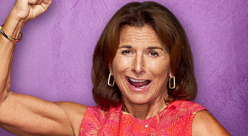 Auf dem Bild zu The 50 Staffel 2 ist Claudia Obert vor einem violetten Hintergrund zu sehen. Sie trägt ein ärmelloses, pink-orange gemustertes Kleid und goldene Ohrringe. Mit einem breiten Lächeln und erhobener Faust wirkt sie energiegeladen und selbstbewusst. Ihre offene Körperhaltung und der lebendige Ausdruck unterstreichen ihre dynamische Ausstrahlung.