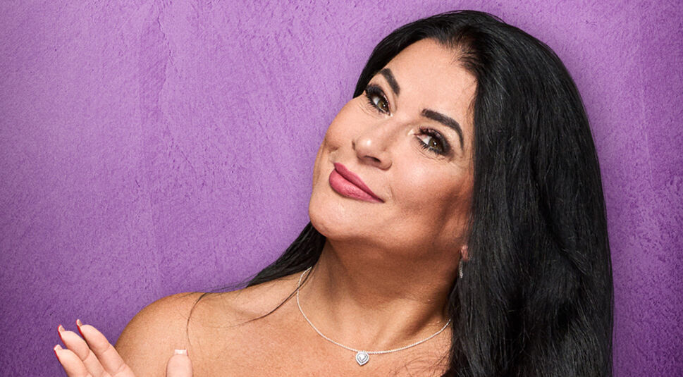 Auf dem Bild zu The 50 Staffel 2 ist Iris Klein vor einem violetten Hintergrund zu sehen. Sie trägt eine silberne Halskette mit Anhänger und hat ihre langen, dunklen Haare offen. Mit einem leichten Lächeln und hochgezogenen Augenbrauen blickt sie charmant in die Kamera. Ihre Handbewegung wirkt verspielt und unterstreicht ihre selbstbewusste Ausstrahlung.