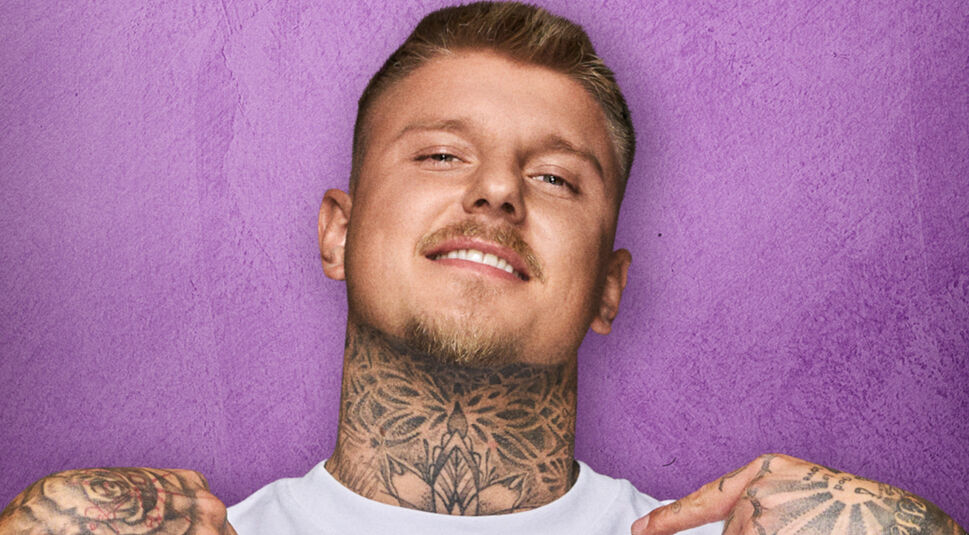 Auf dem Bild zu The 50 Staffel 2 ist Paco Herb vor einem violetten Hintergrund zu sehen. Er trägt ein weißes T-Shirt und zeigt mit beiden Händen auf sich selbst. Sein Hals und seine Hände sind mit auffälligen Tattoos verziert. Paco Herb hat einen selbstbewussten Gesichtsausdruck mit einem leichten Lächeln. Die Nahaufnahme betont seine markanten Gesichtszüge.