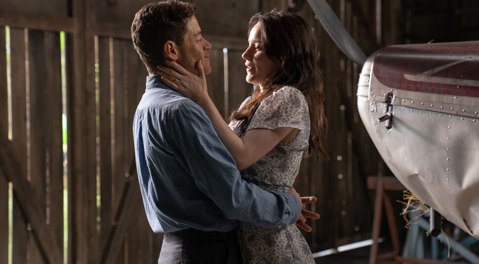 In dieser Szene aus The Amateur sind Charles Heller, gespielt von Rami Malek, und seine Frau Sarah, dargestellt von Rachel Brosnahan, in einer innigen Umarmung zu sehen. Sie stehen in einer rustikalen Scheune neben einem kleinen Flugzeug. Sarah hält zärtlich sein Gesicht, während Charles sie liebevoll anschaut. Die Szene vermittelt emotionale Nähe und einen Moment des Innehaltens.