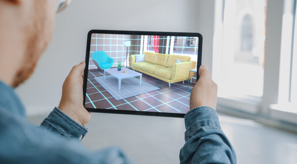 Das Bild zum Thema Was ist Augmented Reality zeigt eine Person, die ein Tablet vor sich hält. Auf dem Display ist ein virtuell eingerichtetes Wohnzimmer mit Sofa, Sessel, Tisch und Lampe zu sehen, das in den realen Raum projiziert wird. Die Szene demonstriert anschaulich, wie Augmented Reality zur Wohnraumplanung genutzt wird.