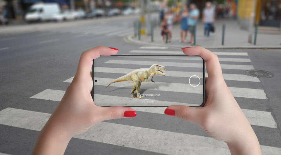 Auf dem Bild zum Thema Was ist Augmented Reality hält eine Person ein Smartphone über einen Zebrastreifen in der Stadt. Auf dem Display erscheint ein digital eingefügter Tyrannosaurus, der scheinbar über die Straße läuft. Die Szene veranschaulicht eindrucksvoll, wie AR digitale Objekte realitätsnah in die echte Umgebung integriert.