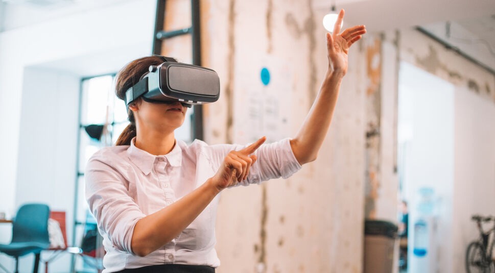 Auf dem Bild zum Thema Was ist Extended Reality sitzt eine Frau mit Virtual-Reality-Brille in einem modernen Büro. Sie hebt die Arme und interagiert mit unsichtbaren virtuellen Inhalten. Die Szene zeigt, wie Extended Reality neue Formen der digitalen Interaktion ermöglicht, selbst in alltäglichen Arbeitsumgebungen.