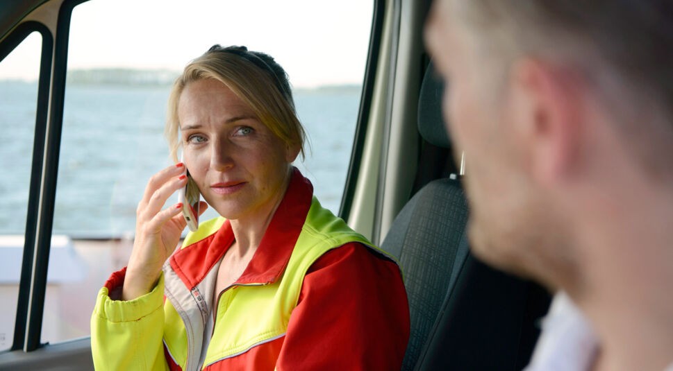 Auf dem Bild aus der ARD-Serie Praxis mit Meerblick, Teil der ARD-Serien 2025, ist Nora Kaminski, gespielt von Tanja Wedhorn, in einem Rettungsfahrzeug zu sehen. Sie trägt eine auffällige rot-gelbe Einsatzjacke und telefoniert mit konzentriertem Blick. Im Vordergrund ist unscharf ein männlicher Gesprächspartner angedeutet. Die Stimmung ist angespannt und ernst.