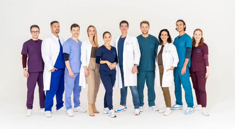 Auf dem Bild zur Serie In aller Freundschaft – Die jungen Ärzte, Teil der ARD-Serien 2025, steht das junge Klinikteam in farblich abgestimmter medizinischer Kleidung vor hellem Hintergrund. Die Darstellerinnen und Darsteller lächeln selbstbewusst in die Kamera und strahlen Teamgeist und Kompetenz aus. Die Szene vermittelt eine moderne und dynamische Krankenhausatmosphäre.