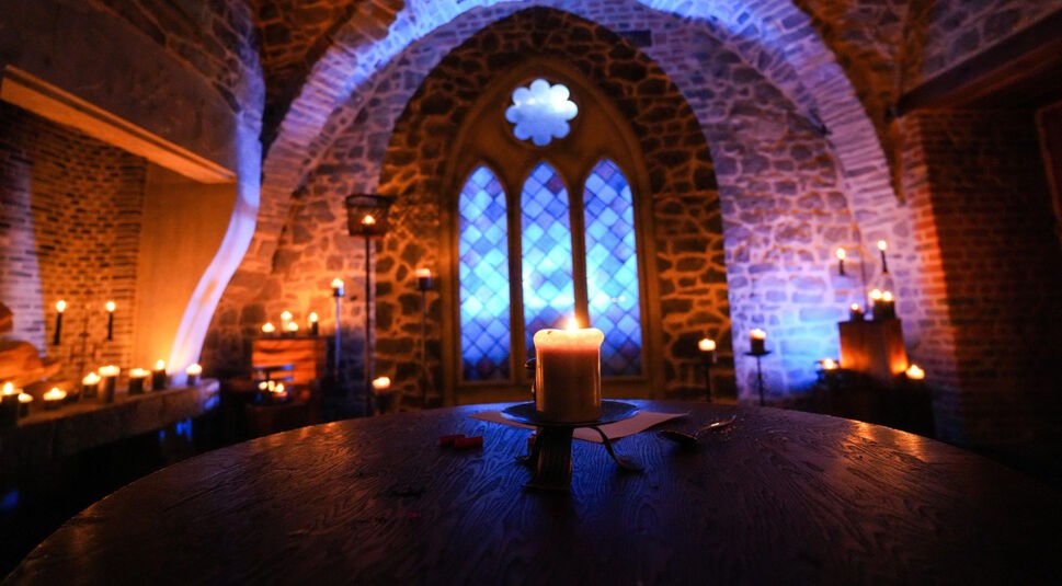 Das Bild zeigt einen geheimnisvoll beleuchteten Raum im Schloss mit groben Steinmauern, Fenstern und dutzenden brennenden Kerzen. Im Vordergrund steht eine einzelne Kerze auf einem runden Holztisch, umgeben von dunkler Atmosphäre. Die Szene wirkt mittelalterlich und feierlich und stammt aus der RTL-Show „Die Verräter - Vertraue Niemandem!“.