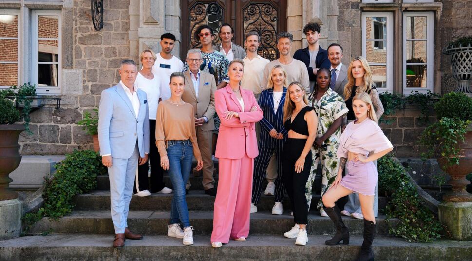 Das Gruppenbild zeigt die prominenten Teilnehmer:innen der dritten Staffel der RTL-Show „Die Verräter - Vertraue Niemandem!“. Vor einem historischen Steingebäude posieren 17 Personen in moderner und stilvoller Kleidung. Im Zentrum steht Moderatorin Sonja Zietlow in einem auffälligen pinken Anzug mit verschränkten Armen.