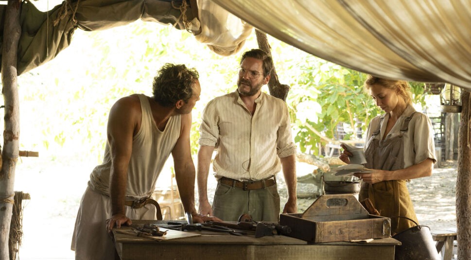 Auf dem Bild zum Thema Eden Ende erklärt sind Jude Law als Dr. Friedrich Ritter, Daniel Brühl als Heinz Wittmer und Vanessa Kirby als Dora Strauch in einer angespannten Szene zu sehen. Sie stehen unter einem provisorischen Zeltdach im Dschungel, umgeben von Werkzeug und Papier. Die Stimmung wirkt ernst und nachdenklich, was das Ende von Eden vorbereitet.