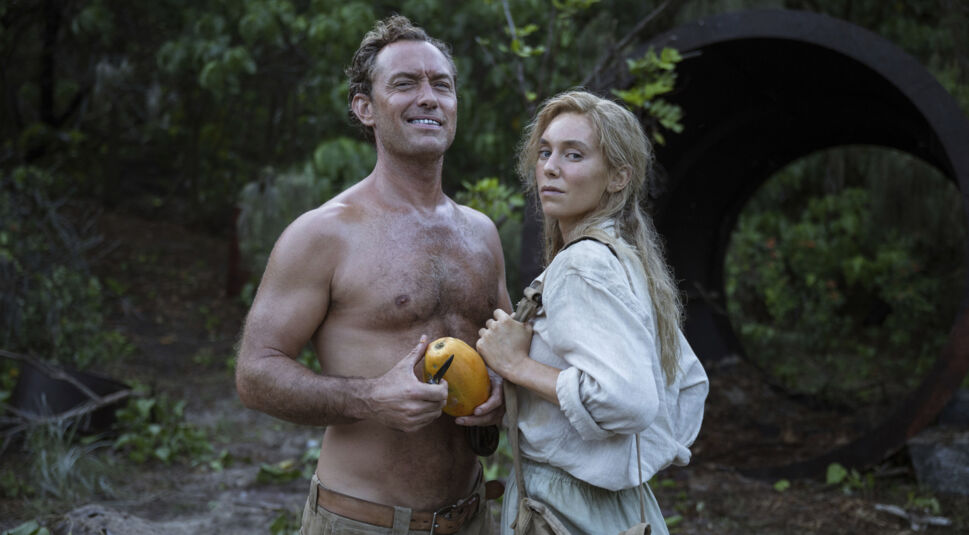 Auf diesem Bild aus dem Film Eden, der auf einer wahren Geschichte basiert, sind Jude Law als Dr. Friedrich Ritter und Vanessa Kirby als Dora Strauch in einer intensiven Szene zu sehen. Die Darstellung fängt die raue Natur und die emotionale Spannung ein: Law steht oberkörperfrei mit einer Papaya in der Hand, während Kirby ihn entschlossen ansieht – beide umgeben von dichter tropischer Vegetation.