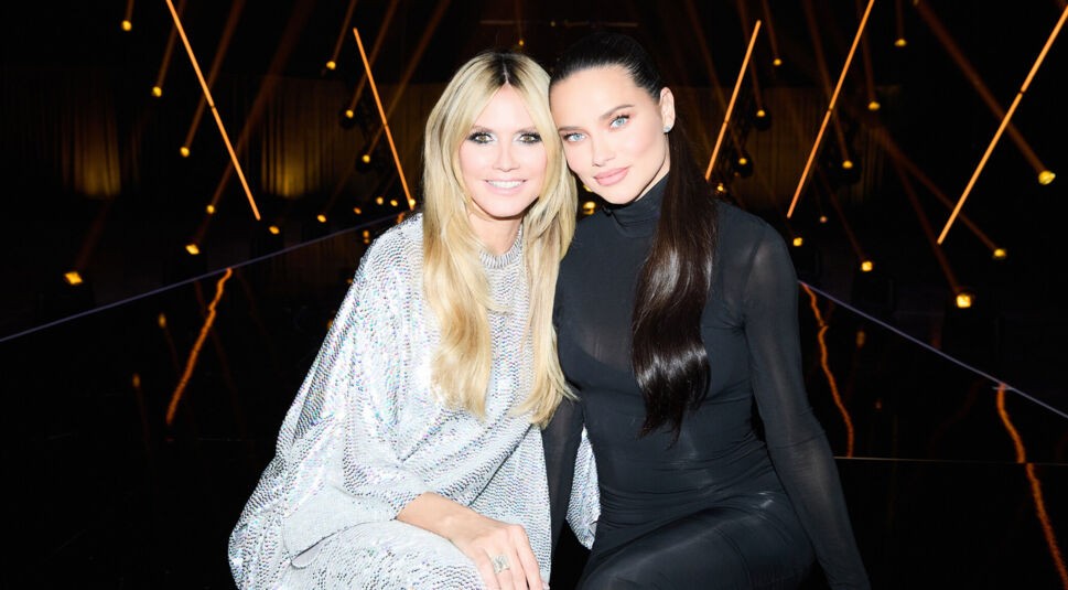 Auf diesem Bild aus GNTM 2025 Folge 14 sind Heidi Klum und Adriana Lima nebeneinander auf dem Laufsteg zu sehen. Heidi Klum trägt ein funkelndes silbernes Outfit und lächelt direkt in die Kamera, während Adriana Lima in einem eleganten, schwarzen, figurbetonten Look daneben sitzt. Der Hintergrund ist mit dramatischer Lichtinszenierung versehen und vermittelt eine glamouröse Show-Atmosphäre.