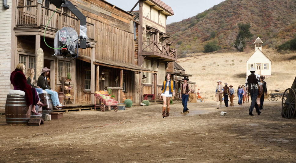 In dieser Szene aus GNTM 2025 Folge 15 betreten mehrere Kandidat:innen in Western-Outfits ein Set. Im Vordergrund läuft eine Frau in gelber Weste und weißen Shorts über den staubigen Boden. Außerdem sieht man eine hölzerne Häuserzeile, ein Western-Gebäude auf einem Hügel und eine Filmkamera.