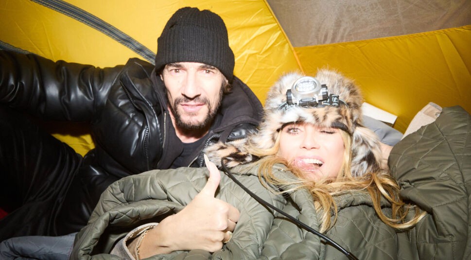 Im Bild zur GNTM 2025 Folge 16 sind Thomas Hayo und Heidi Klum in einem gelben Zelt zu sehen. Thomas trägt eine schwarze Mütze und eine dicke Lederjacke, während Heidi eine Pelzmütze mit Stirnlampe trägt und mit einem Daumen nach oben fröhlich in die Kamera blickt.