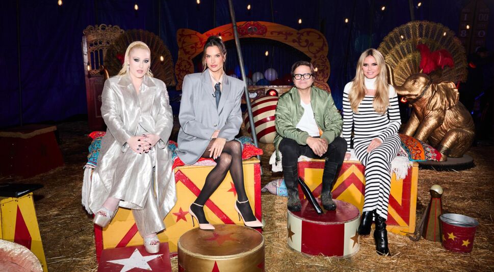 Auf dem Bild zur GNTM 2025 Folge 17 sitzen die vier Jurymitglieder Hayley Hasselhoff, Alessandra Ambrosio, Kristian Schuller und Heidi Klum in einem aufwendig gestalteten Zirkus-Setting. Hayley trägt ein glänzendes silbernes Outfit, Alessandra ein oversized graues Sakko mit Krawatte, Kristian Schuller eine grüne Jacke und Heidi ein gestreiftes Schwarz-Weiß-Kleid. Die Szenerie ist farbenfroh, mit Zirkusrequisiten, Stroh und einem goldenen Elefanten im Hintergrund.