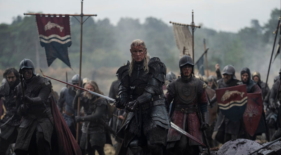 Auf dem Bild zu House of the Dragon Staffel 3 ist Matt Smith als Daemon Targaryen in schwarzer, kampferprobter Rüstung mit gezogenem Schwert zu sehen. Er steht entschlossen an vorderster Front eines mittelalterlichen Schlachtfelds, umgeben von schwer gerüsteten Soldaten mit blutverschmierten Gesichtern. Die Szene wirkt düster, angespannt und voller Dramatik.