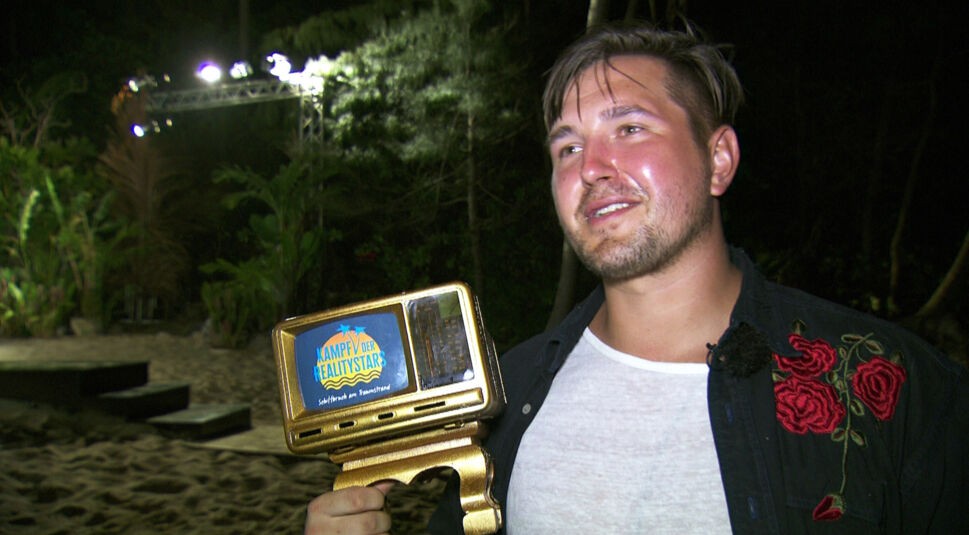 Auf dem Bild zum Thema Kampf der Realitystars-Gewinner:innen ist Kevin Pannewitz in einem nächtlich beleuchteten Setting zu sehen. Er lächelt zufrieden und hält die goldene Sieger-Trophäe der Show in der Hand. Kevin trägt ein schwarzes Hemd mit auffälligem Rosenmuster über einem weißen Shirt.