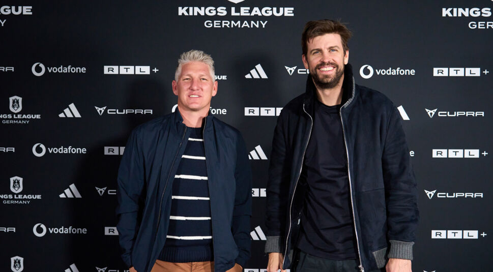 Auf dem Bild zur Kings League Germany posieren Bastian Schweinsteiger und Gerard Piqué nebeneinander vor einer Sponsorenwand. Beide tragen dunkle Jacken, lächeln in die Kamera und wirken entspannt und kollegial. Im Hintergrund sind Logos der Kings League Germany, RTL+, Vodafone, Adidas und Cupra zu sehen – passend zum offiziellen Start des neuen Fußballformats in Deutschland.
